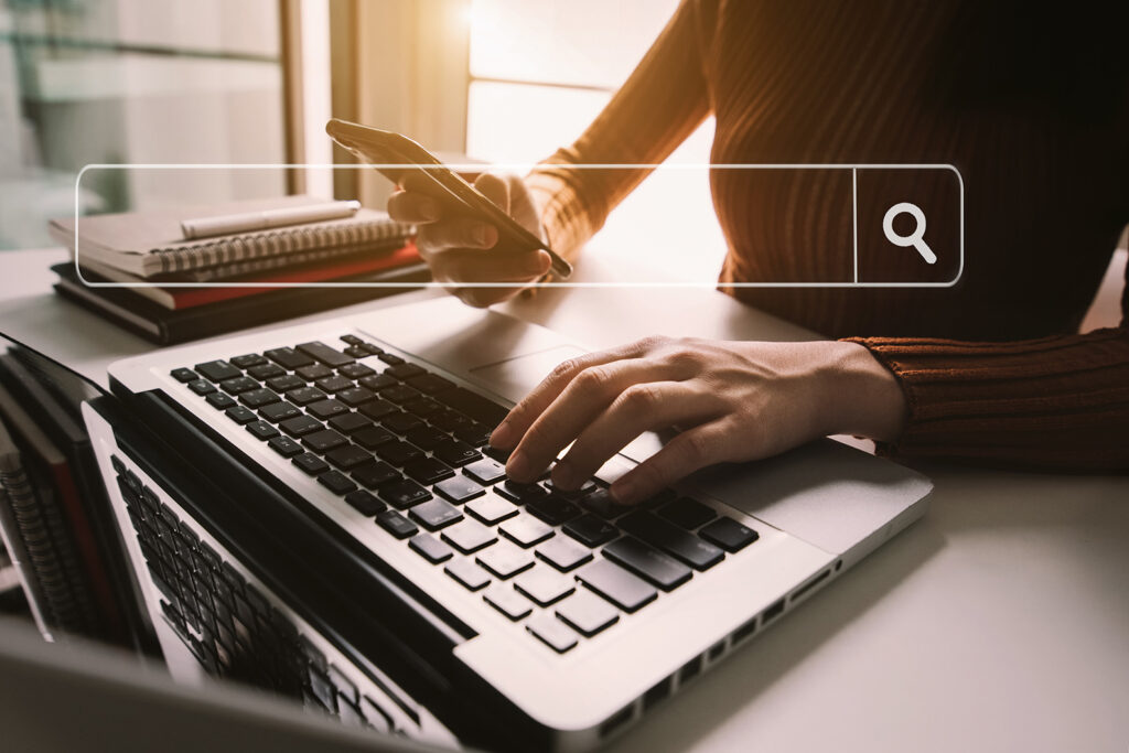A person typing on a laptop with one hand while holding a smartphone with the other, with a graphic overlay of a search bar and magnifying glass icon.