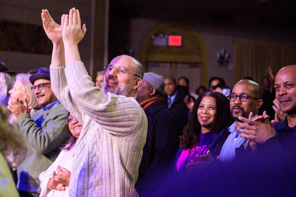 A diverse group of people in a dimly lit room applaud enthusiastically, with smiles and a sense of joy. The atmosphere is lively and celebratory.